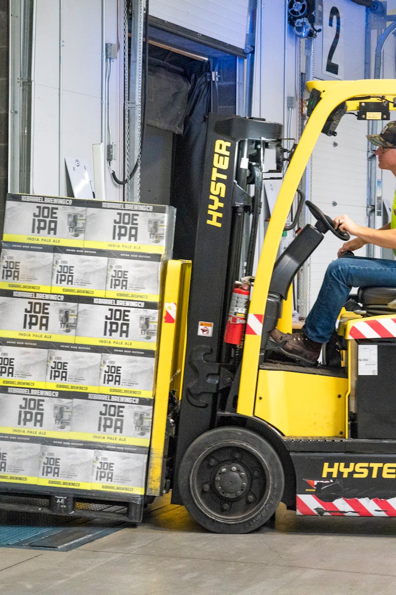 A worker drives a Hyster forklift moving Joe IPA boxes in a warehouse.